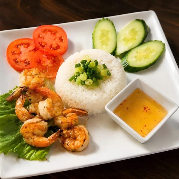 Grilled shrimp, white rice with green onions, sliced tomatoes, cucumbers, and dipping sauce on a white plate.