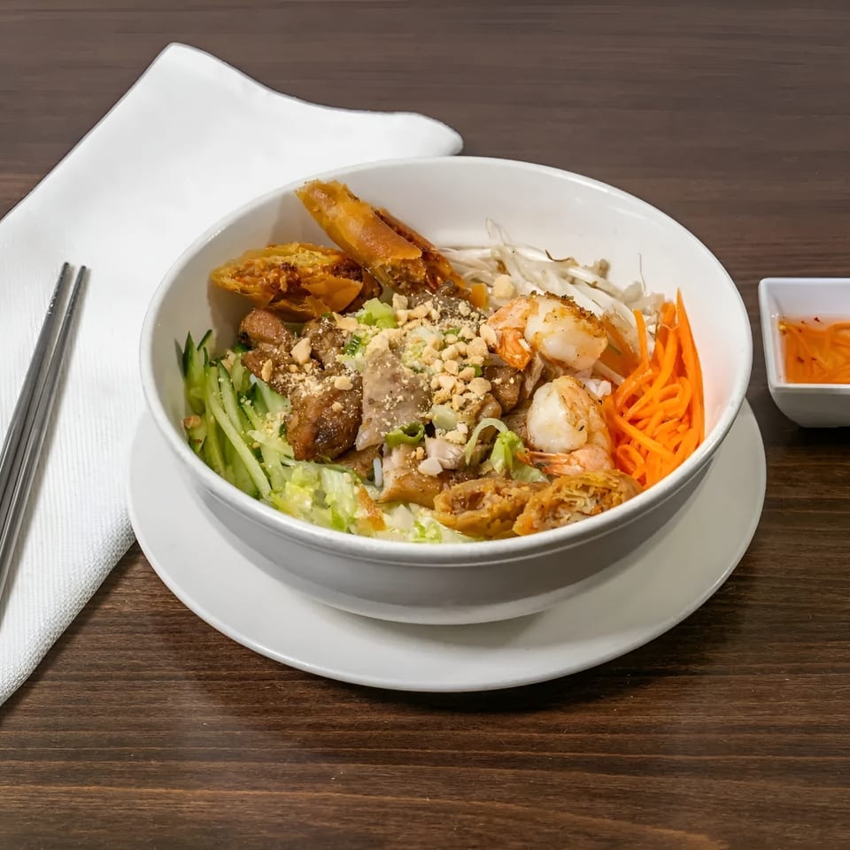 A Vietnamese vermicelli bowl with shrimp, pork, egg rolls, and vegetables, served with sauce.