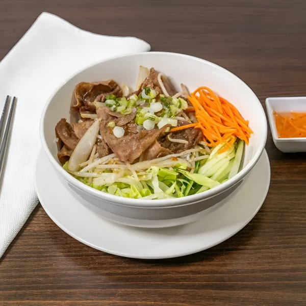 A white bowl of Vietnamese vermicelli with grilled pork, fresh vegetables, and a side of sauce.
